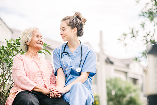 Young nurse in blue scrubs and wearing stethoscope sitting next to elderly asian woman outside